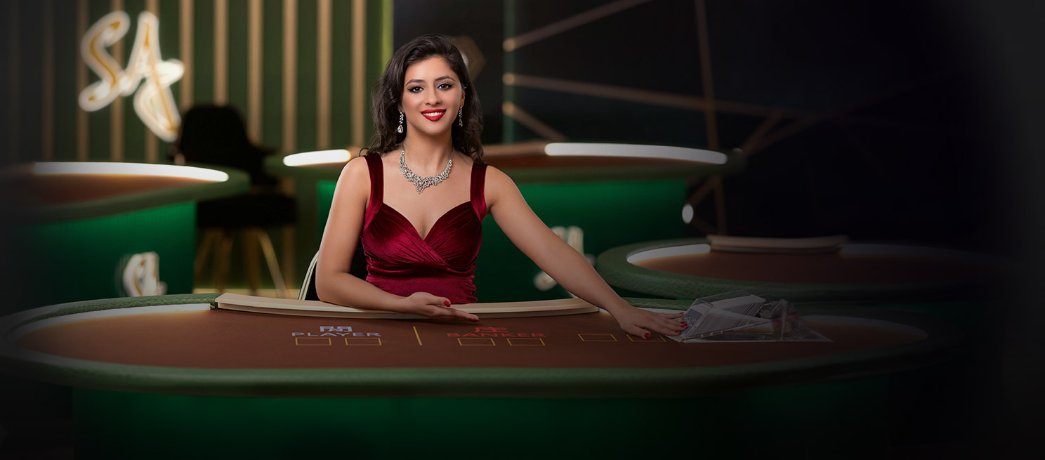 Smiling woman in a red dress seated at a casino table, elegantly presenting playing cards, with gaming tables in the background.