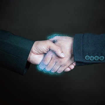 Close-up of two hands shaking, one in a black suit and the other in a grey suit. The handshake is partially represented with a glowing, digital effect, symbolizing partnership and collaboration in a modern context.